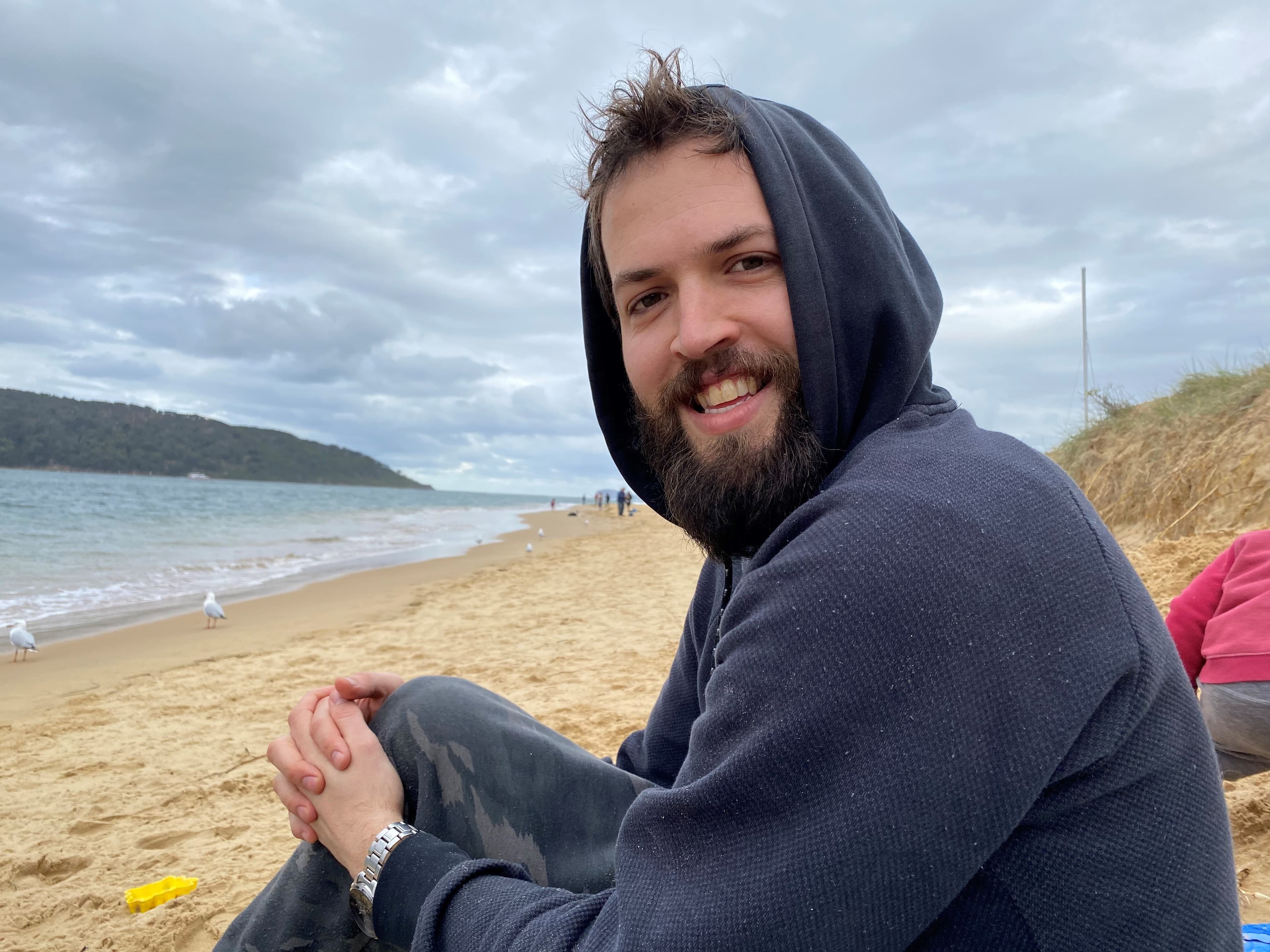 Silas, a man living with multiple sclerosis, is wearing a dark blue hoodie and is seated on the beach.