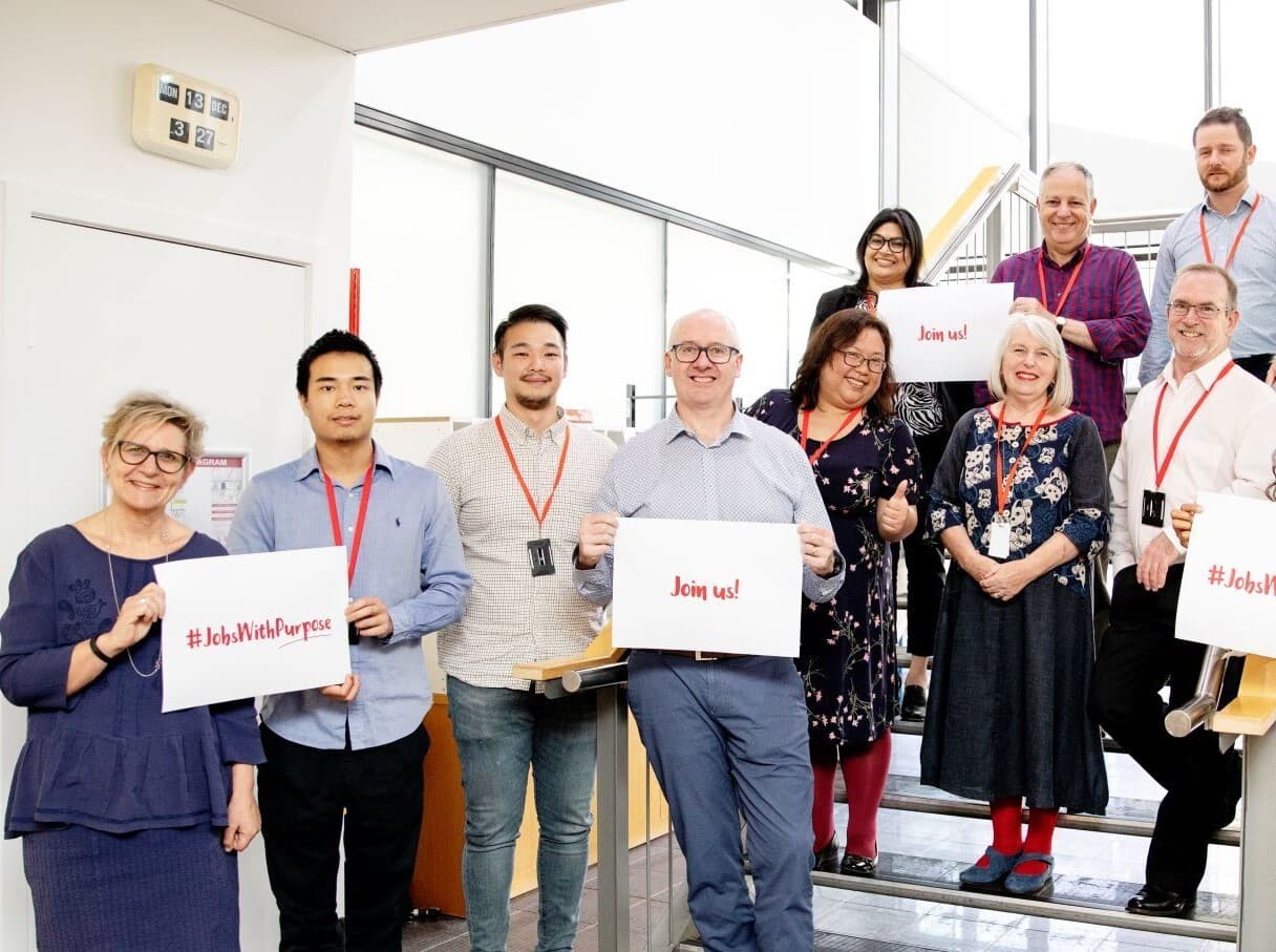 A group of MS Plus staff members standing on the stairs and holding up cards with messages to encourage people to work for MS Plus.