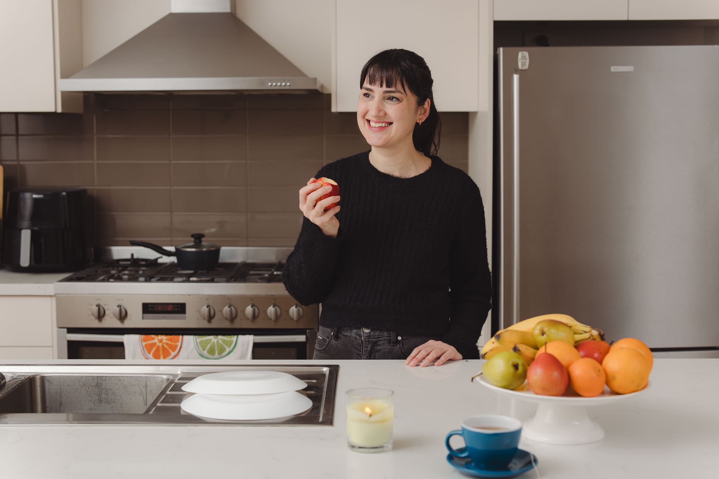 A woman smiling while standing in kitchen holding and eating an apple