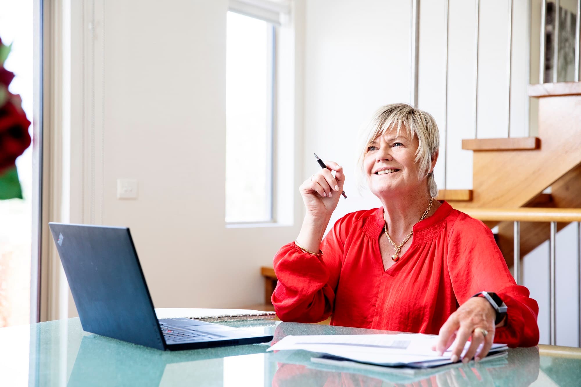 A woman sitting and working on laptop holding a pen with another hand resting on notepad.